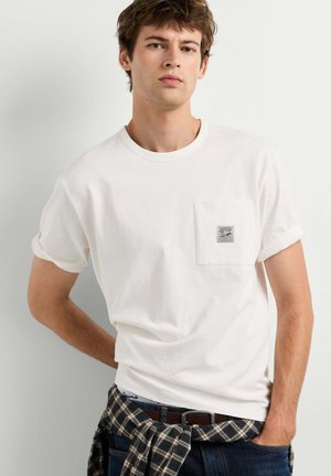 Young man with short brown hair wears a white t-shirt with a chest pocket and a rolled-up sleeve, dark jeans, and a plaid shirt tied around his waist.