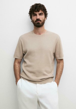 Baardige man met krullend haar, gekleed in een beige textuur korte mouw trui en witte broek, staand met handen in de zakken tegen een witte muur.