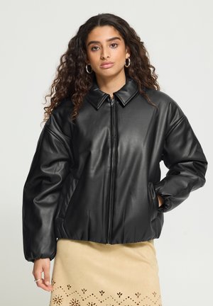Femme aux longs cheveux bouclés porte une veste en cuir noire zippée et une jupe beige avec des motifs découpés, debout avec une main dans la poche.