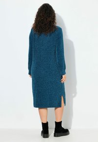 Abito in maglia color teal con maniche lunghe e spacchi laterali. Presenta un tessuto testurizzato e arriva sotto il ginocchio. Indossato con stivaletti neri alla caviglia.