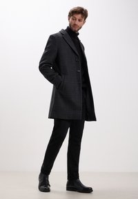 Grau karierter Mantel mit taillierter Passform, mit Reverskragen, drei Knöpfen und seitlichen Taschen, kombiniert mit schwarzen Hosen und Leder-Stiefeletten.