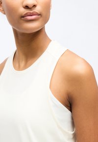 Κοντινό πλάνο ενός ατόμου που φοράει αμάνικη λευκή αθλητική μπλούζα, εμφανίζοντας τον λαιμό, τον ώμο και το επάνω μέρος του στήθους έναντι ενός απλού φόντου.