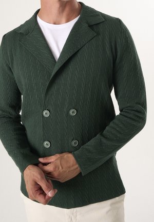 Hombre con blazer de doble botonadura texturizado de color verde oscuro sobre camisa blanca, ajustándose el blazer con las manos, con fondo liso y claro.