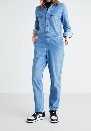 Personne portant une combinaison en jean bleu clair avec les manches retroussées et des baskets noir et blanc, debout les mains dans les poches.