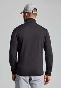 Ein Mann trägt eine schwarze strukturierte Jacke mit langen Ärmeln, hellgraue Hose und eine hellgraue Baseballkappe und blickt von der Kamera weg.