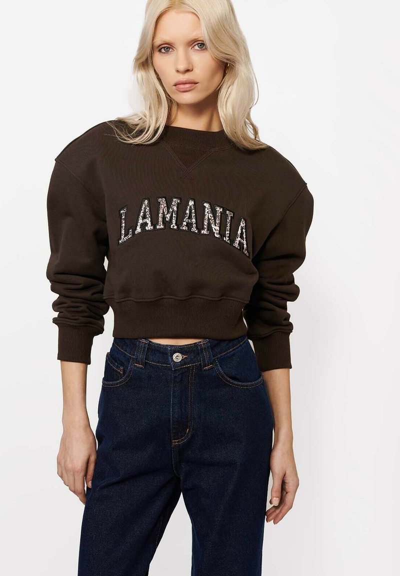 Femeie blondă purtând un sweatshirt scurt maro închis cu textul „LAMANIA” și blugi cu talie înaltă, de culoare albastru închis, pe un fundal alb.