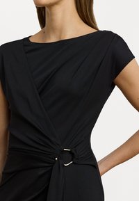 Zwarte jurk met een gedrapeerde halslijn, kapmouwen en een gedraaide taille-detail met een zilveren ringaccent. Gladde, rekbare stof.