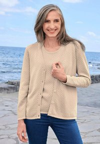 Beiger Strickcardigan mit strukturiertem Bobbelmuster, Rundhalsausschnitt, Knopfverschluss und gewellten Kanten, kombiniert mit einem passenden Oberteil und blauen Jeans.