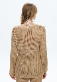 Woman with blonde hair wearing beige open-knit long-sleeve top and matching pants, shown from the back against a plain white background.