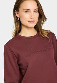 Bordový svetri s okrúhlym výstrihom, dlhými rukávmi a vyšitým nápisom "Smile" na prednej strane. Mäkká tkanina s hladkou textúrou.