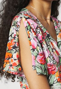 Femme portant un haut sans manches à fleurs avec des manches fendue et un fin collier en argent, avec des cheveux bouclés et foncés visibles.