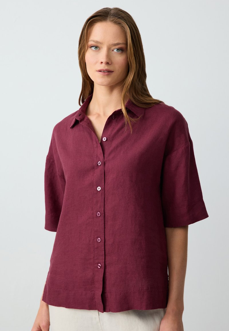Camicia di lino bordeaux con bottoni davanti, maniche corte e colletto. Presenta bottoni viola e una vestibilità rilassata. Tessuto morbido.