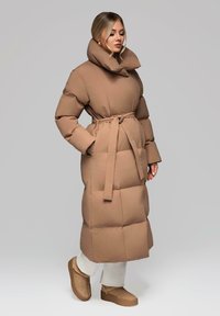 Žena v dlouhém hnědém vatovaném kabátu s vysokým límcem a páskem, bílých kalhotách a odpovídajících hnědých kotníkových botách, stojící na jednobarevném pozadí.