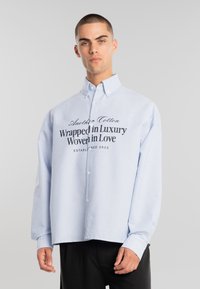 Joven con cabello corto que lleva una camisa de botones de rayas azul claro con el texto "Envuelto en lujo, tejido con amor" y pantalones negros.