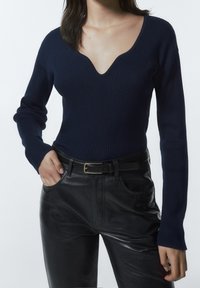 Marinblå ribbstickad tröja med djup V-ringning och långa ärmar, kombinerad med svarta läderbyxor och ett svart bälte med en guldfärgad spänne.