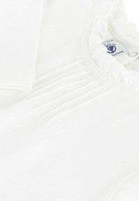 Weißes Kleidungsstück mit gerafftem Halsausschnitt und Falten. Weicher Stoff, der ein Pflegeetikett aufweist. Keine sichtbaren Muster.