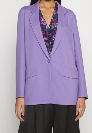 Kvinde iført en lavendel blazer over en blomsterprintet lilla bluse og sorte bukser med vide ben, stående med armene afslappet ned langs siden.