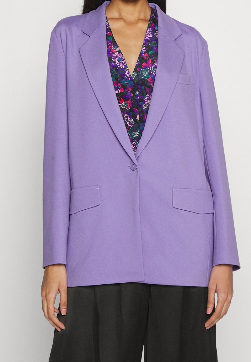 Femme portant un blazer lavande sur un chemisier violet à motifs floraux et un pantalon large noir, debout avec les bras détendus le long du corps.
