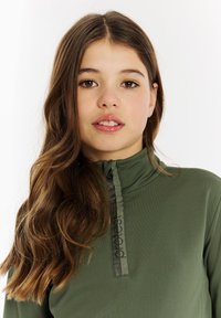 Groene zip-up pullover met hoge kraag, voorzien van een textuurstof en een rits aan de voorkant met een logo-accent. Lang haar gedeeltelijk zichtbaar.