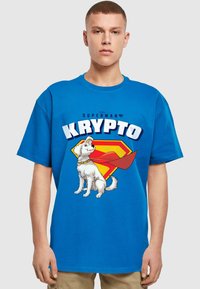 Mėlynas medvilninis marškinėliai su balto šuns Krypto, dėvinčio apsiaustą ir kaklaraištį, grafika, šalia geltonos ir raudonos „Supermano“ logotipo.