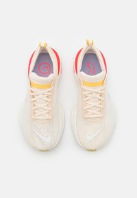 Paire de chaussures de course Nike beige avec logos swoosh blancs, accents orange au talon, languettes jaunes et semelles intérieures violettes sur fond blanc.