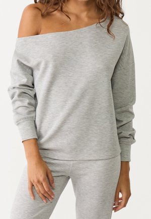 Femme portant un top gris clair à épaules dénudées et manches longues avec un pantalon assorti, montrant une tenue décontractée du cou aux hanches.