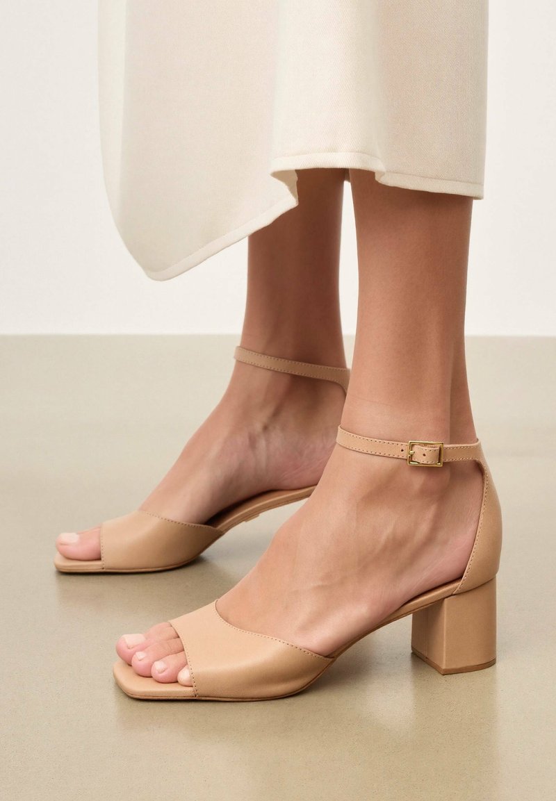 Close-up of feet wearing beige open-toe block heel sandals with ankle straps, paired with off-white cropped pants.