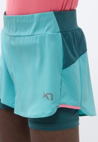 Αθλητικό σορτς σε γαλάζιο του νερού με γαλάζια-πράσινη ζώνη μέσης, με διάτρητη υφή και ροζ λεπτομέρεια. Το λογότυπο είναι ορατό στο κάτω αριστερό μέρος.