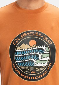 Orange cotton t-shirt featuring a circular graphic with waves, mountains, and sun. Text reads "Quiksilver Boardriding Co." in bold.