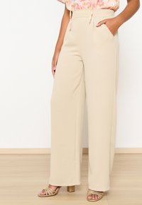 Beige wijde broek van lichtgewicht stof, met een hoge tailleband en zijzakken, gedragen met gouden sandaaltjes met bandjes.