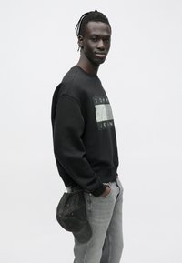 Hombre con rastas que lleva una sudadera negra de Tommy Jeans y vaqueros grises, sosteniendo una gorra de mezclilla oscura detrás de su espalda.