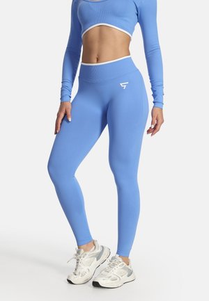 Frau trägt ein blaues, langärmeliges Crop-Top und passende hoch taillierte Leggings, kombiniert mit weißen Sportschuhen, stehend vor weißem Hintergrund.