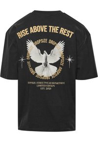 Sort bomulds-t-shirt med en grafisk hvid due. Teksten "RISE ABOVE THE REST" samt brandoplysninger i guld over og under billedet.