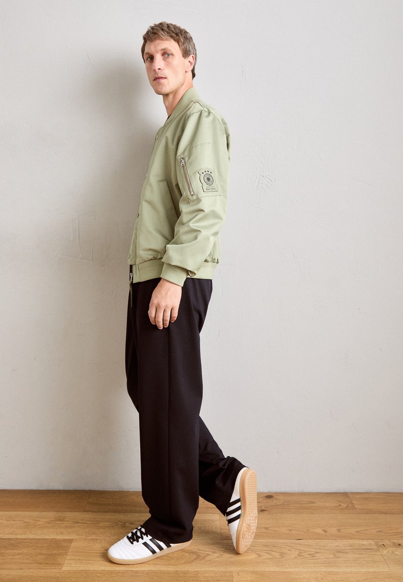 Homme de profil portant une veste bombardier olive, un pantalon noir et des baskets blanches avec des rayures noires, debout sur un sol en bois devant un mur uni.