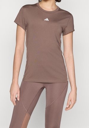 Femme portant un t-shirt de sport marron ajusté et un legging assorti, debout devant un fond uni.