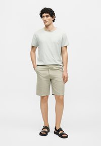 Homme aux cheveux bouclés portant un t-shirt gris clair, un short beige et des sandales noires, debout avec une main dans la poche devant un fond uni.