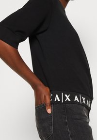 Černá mikina s krátkými rukávy, volným střihem a kontrastním bílým žebrovaným lemem s logem. Materiál má hladkou strukturu.