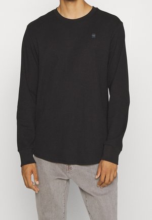 Homme portant un t-shirt noir uni à manches longues côtelé et un jean gris clair, debout les mains détendues le long du corps sur un fond blanc.