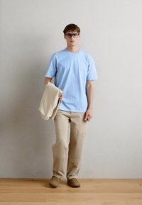 Ανοιχτό μπλε βαμβακερό t-shirt, μπεζ παντελόνι σε χαλαρή γραμμή, κρατώντας ένα μπεζ φούτερ, καφέ χρωματιστά παπούτσια χωρίς πέλμα, μινιμαλιστικό φόντο.