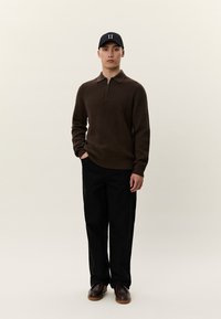 Vyras dėvi juodą beisbolo kepuraitę, rudos spalvos ketvirtinio užtrauktuko megztinį, juodas kelnes ir rudos spalvos batus, stovi priešais šviesią, vienspalvę foną.