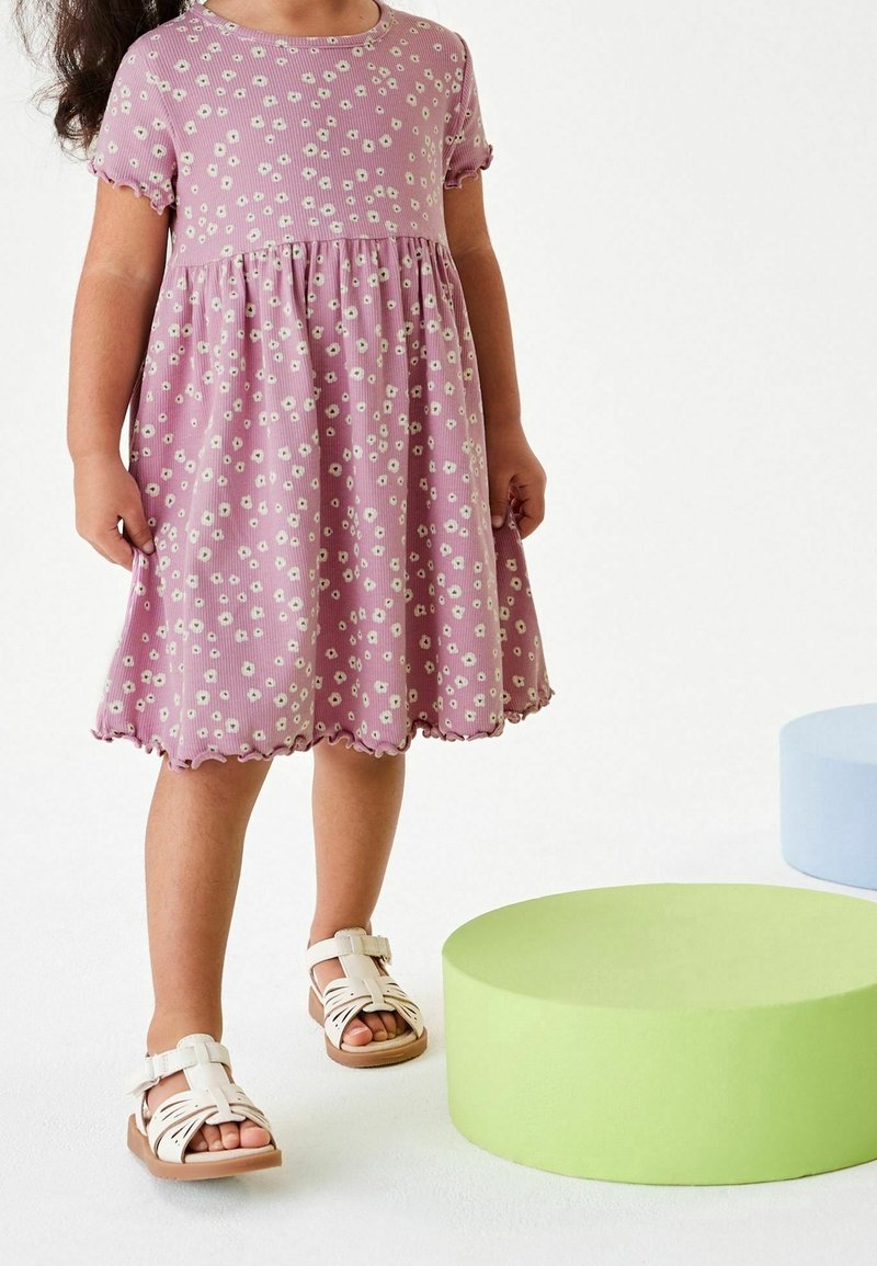 Enfant portant une robe violette avec un motif floral blanc et des sandales blanches, debout près de coussins ronds verts et bleus sur un sol blanc.