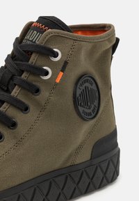Botine din pânză verde măsliniu, la glezna, cu șireturi negre și talpă din cauciuc. Include un patch rotund cu logo negru și cusături contrastante portocalii.