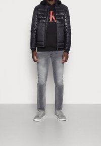 Chaqueta negra acolchada sobre una sudadera negra con capucha, jeans grises y zapatillas grises; cuenta con cremalleras, logo y una textura elegante y suave.