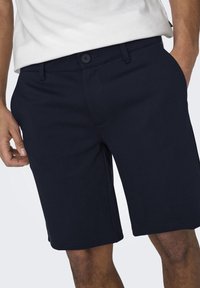 Hombre vestido con pantalones cortos oscuros de color marino abotonados y una camisa blanca, con una mano en el bolsillo izquierdo y la otra mano relajada.