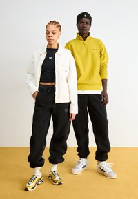 Biely sako, čierny crop top a čierne fleecové nohavice nosí jedna osoba; žltá mikina a čierne nohavice sú oblečené druhou osobou. Obuv v zlatej a sivej farbe.