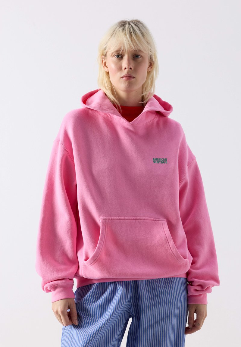 Naine kannab üle suuruse heledat roosat kapuutsiga dressipluusi, millel on ees tasku ja tekst "American Vintage", ning lahtisi sinise ja valge triibulisi pükse valge tausta ees.