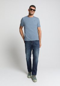 Lichtblauwe katoenen T-shirt met korte mouwen, gecombineerd met donkere spijkerbroeken. Model draagt grijze wandelschoenen en een zonnebril. Eenvoudig ontwerp, casual pasvorm.