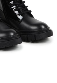 Botas de cuero negro hasta el tobillo con suelas texturizadas, cierres con cordones y acentos de charol brillantes en los laterales. Diseño robusto y construcción sólida.