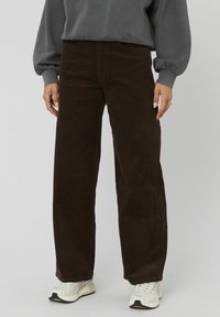 Pantalon en velours côtelé marron avec une coupe large, présentant un ribbing vertical et des poches avant. Assorti à un sweat en gris et des chaussures de sport.