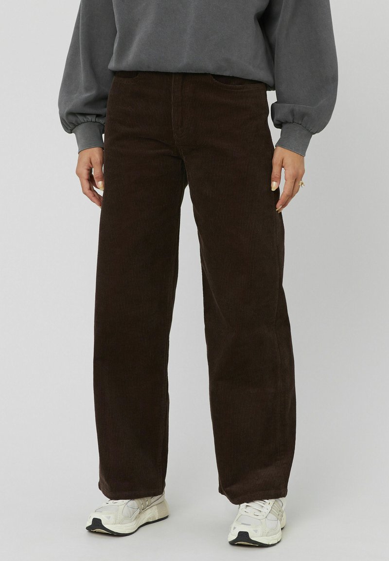 Pantalon en velours côtelé marron avec une coupe large, présentant un ribbing vertical et des poches avant. Assorti à un sweat en gris et des chaussures de sport.